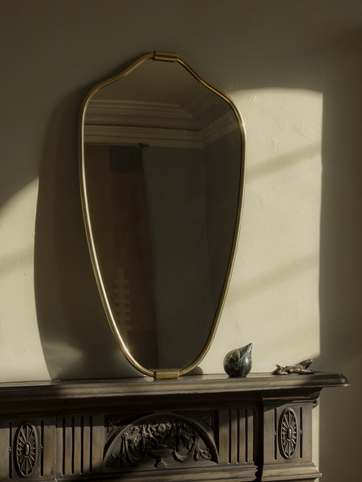 Senne Mirror in brass on a dark carved mantel with late sunlight and shadow across the wall
