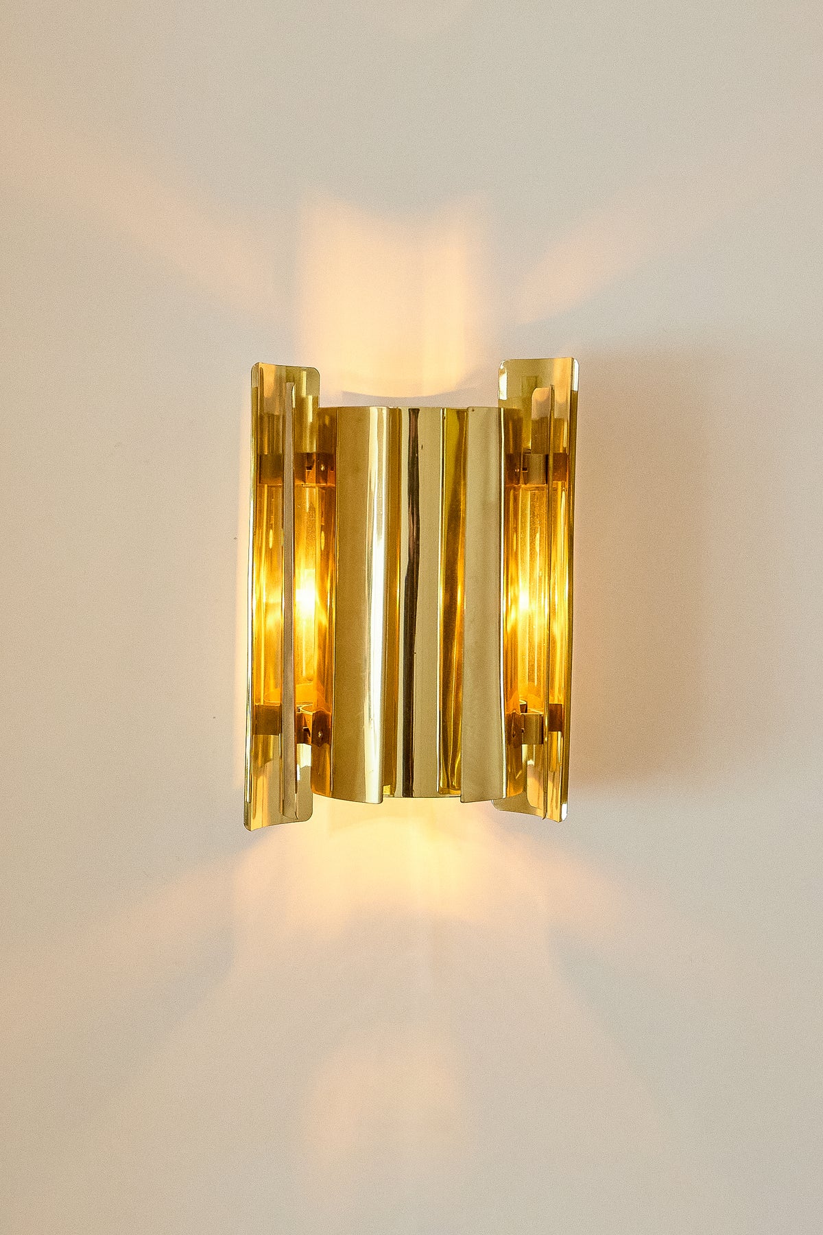 Falkenbergs Belysning brass sconce, front view, lit, with the brass sheets overlapping